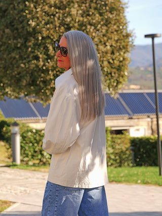 Eine silberhaarige Frau trägt die OFF#DLY Jacke Patch creme mit dekorativen Brust-Patches, leichtem Beige, klassischem Kragen und lässigem Schnitt, während sie in einem sonnigen Garten steht.