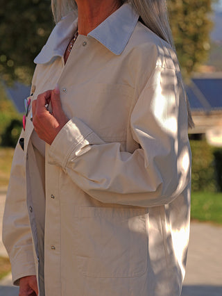 Person trägt die OFF#DLY Jacke Patch creme, einen knielangen, hellbeigen Mantel mit blauem Kragen, Snap-Verschlüssen und aufgesetzten Taschen, der lässig und modern wirkt.