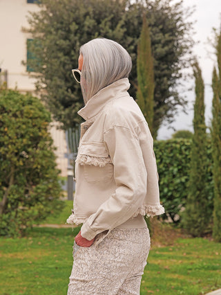 Person im Profil trägt die OFF#DLY Denimjacke Dana beige mit modischen Fransen an Brust und Saum, kombiniert mit hochgeschnittenen, gemusterten Hosen, in einem Garten mit Bäumen im Hintergrund.