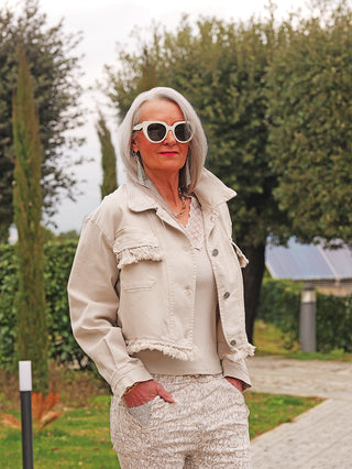 Frau trägt die OFF#DLY Denimjacke Dana beige mit Fransen, kurzen Schnitt und Brusttaschen. Kombiniert mit einem Spitzenoberteil und gemusterten Hosen auf einem Gartenweg.