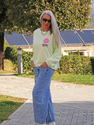 Frau im Garten trägt den „OFF#DLY Sweat Happy mint“, einen lässigen, mintfarbenen Pullover mit Smiley-Print, kombiniert mit blauer Jeans und weißen Sneakers, vermittelt entspannten, fröhlichen Stil.