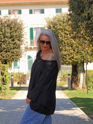 Frau trägt das OFF#DLY Shirt Garda black mit Off-Shoulder-Ausschnitt und ausgestellten Ärmeln, kombiniert mit einer Silberkette, vor einer gartenartigen Kulisse.