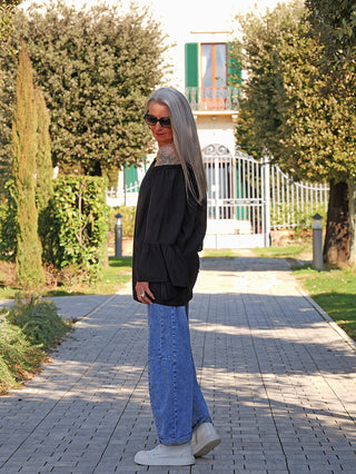 Person trägt das OFF#DLY Shirt Garda black mit off-shoulder Ausschnitt und weiten Ärmeln. Das fließende Design ergänzt stilvoll die weit geschnittenen Jeans und weißen Sneakers auf einem Gartenweg.