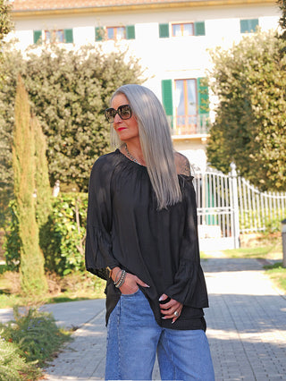 Frau im OFF#DLY Shirt Garda black mit weiten, voluminösen Ärmeln und lockerem Schnitt, kombiniert mit hellen Jeans, steht auf Gehweg vor grünen Pflanzen und einem Gebäude mit grünen Fensterläden.