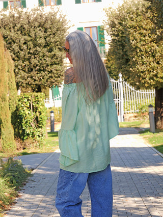 Person mit silbernem Haar trägt das OFF#DLY Shirt Garda mint, ein lockeres, schulterfreies Blusen-Shirt mit Volantärmeln, sichtbar über Jeans, im Garten stehend vor Hecke und Zaun.