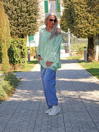 Frau in lockerem, mintgrünem OFF#DLY Shirt Garda mit offener Schulter, ausgestellten Ärmeln und entspannter Passform, kombiniert mit hochgekrempelten Jeans und weißen Sneakers, auf Gartenweg stehend.