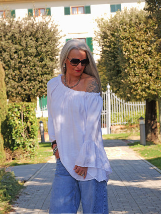 Frau trägt das OFF#DLY Shirt Garda white mit weitem, elastischem Off-Shoulder-Ausschnitt und voluminösen Volant-Ärmeln, kombiniert mit blauen Schlagjeans, in einer sonnigen Gartenlandschaft.