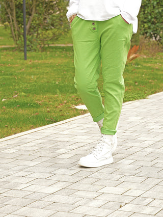 Person trägt OFF#DLY Joggpant Lilly green, hellgrüne Hosen mit elastischem Bund und angedeuteter Knopfleiste, während sie auf einem gepflasterten Weg steht. Kombiniert mit weißen High-Top-Sneakern.