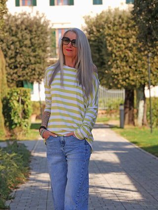 Ältere Frau trägt das OFF#DLY Ringel Grosso white/matcha Langarmshirt mit lockerer Passform und Streifen, kombiniert mit blauer Jeans, auf einem Gartenweg, Hände in den Taschen, stilvoll und bequem.