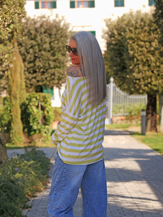 Person trägt das OFF#DLY Ringel Grosso white/matcha, ein locker sitzendes, gelb-weiß gestreiftes Langarmshirt, das leicht über eine Schulter fällt und ein Tattoo enthüllt, in einem sonnigen Garten.