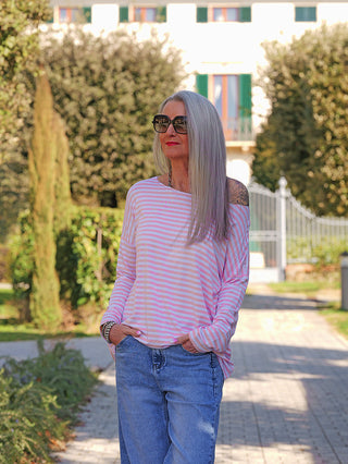 Frau trägt das OFF#DLY Ringelshirt Frida rosa/white, locker und lässig über einer Schulter, kombiniert mit Jeans. Sichtbar: Streifenmuster, entspannte Passform, ideal für stilvolle Freizeitlooks.