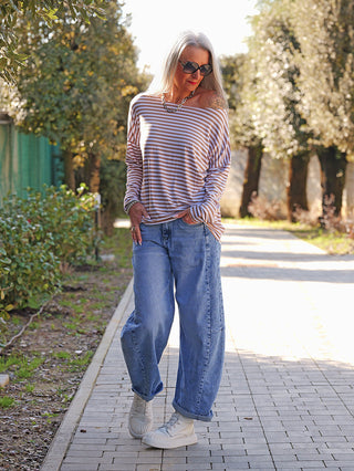 Frau trägt das OFF#DLY Ringelshirt Frida in Weiß/Kamel, kombiniert mit blauen Jeans und weißen Sneakers, während sie einen sonnigen, von Bäumen gesäumten Weg entlanggeht.