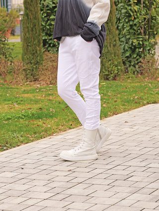 Person trägt OFF#DLY Joggpant Vera white, schmale weiße Hose aus fließendem Stoff, kombiniert mit weißen High-Top-Stiefeln. Bequemer, stilvoller Look auf gepflastertem Weg.