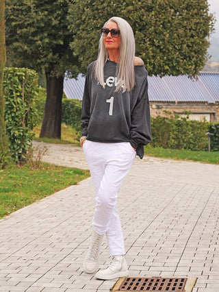 Frau trägt OFF#DLY Joggpant Vera white, weiße Slim-Fit-Hose, kombiniert mit lockerem, off-shoulder Sweatshirt und weißen Sneakers, steht lässig auf Gartenweg.