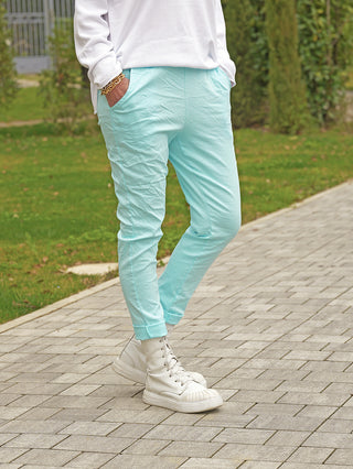 Person trägt OFF#DLY Joggpant Vera türkis, schmal geschnitten mit umgeschlagenem Saum, kombiniert mit weißen klobigen Schnürstiefeln. Sichtbarer goldener Armreif an der Hand in der Tasche.