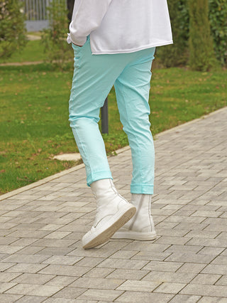 Person trägt OFF#DLY Joggpant Vera türkis, schmal geschnitten, mit weißem Langarmtop und weißen High-Top-Stiefeln, spaziert auf einem Weg neben einem Rasen.