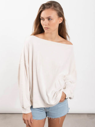 Eine junge Frau trägt das Outryght Cory T chalk, ein locker sitzendes, schulterfreies Sweatshirt aus Cosmo Terry Sweat, kombiniert mit hellen Denim-Cutoff-Shorts.