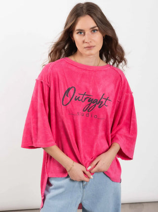 Junge Frau trägt ein Outryght Loo T fuchsia pink Oberteil aus Cosmo Terry Sweat, mit handgeschriebenem Outright studio-Schriftzug, kombiniert mit hellen Jeans oder Shorts, schaut in die Kamera.