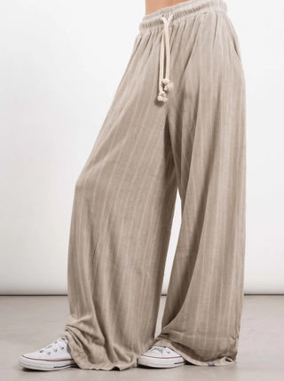 Person trägt Outryght Kamo gravel striped Hose, superweite beige Nadelstreifen mit Kordelzug, die um die Knöchel fallen, kombiniert mit einfachen weißen Low-Top-Sneakers.