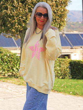 Person trägt den OFF#DLY Sweater Star yellow/pink mit einem übergroßen Schnitt und hot-pinkem Stern-Design, kombiniert mit blauer Jeans, auf einem gepflasterten Weg im Freien.