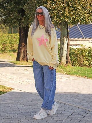 Eine reife Frau trägt den OFF#DLY Sweater Star yellow/pink mit pinkem Stern-Print, kombiniert mit blauer Jeans und weißen Sneakers, während sie auf einem Gehweg spaziert.