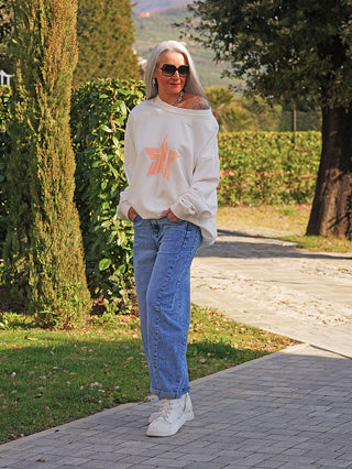 Eine Frau trägt den OFF#DLY Sweater Star white/orange, ein oversized ivory Sweatshirt mit stilisiertem Sternmotiv, kombiniert mit blauen Jeans und weißen High-Top-Sneakers.
