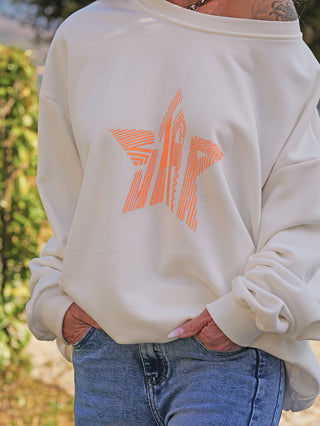 OFF#DLY Sweater Star white/orange: Locker sitzender cremefarbener Pullover mit orangefarbenem geometrischen Stern-Motiv, kombiniert mit blauer Jeans. Hände in den Taschen, moderne Freizeitmode.