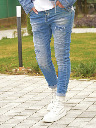 Off#DLY Denim Dertroy San Remo midblue Jeans mit moderner Destroyed-Optik und Used-Waschung, getragen mit weißen High-Top-Stiefeln, perfekt für einen lässigen, urbanen Look auf einem gepflasterten Weg.