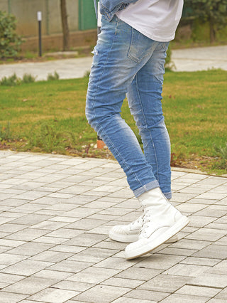 Person trägt OFF#DLY Denim Dertroy San Remo midblue Jeans mit kleinem Riss am Oberschenkel und chunky weißen High-Top-Sneakern, auf gepflastertem Weg, Hintergrund mit Gras und Garten.