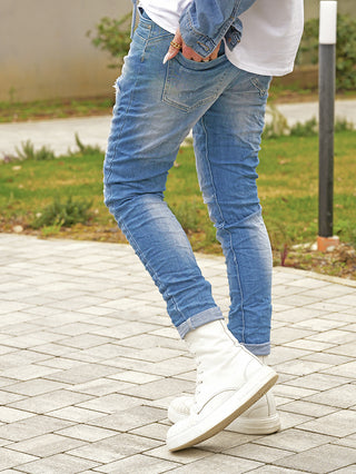 Person trägt OFF#DLY Denim Dertroy San Remo midblue, eine lässige, leicht zerstörte Jeans. Die Passform ist schlank, mit umgeschlagenen Bündchen und einer Hand im Gesäßtasche für einen urbanen Look.