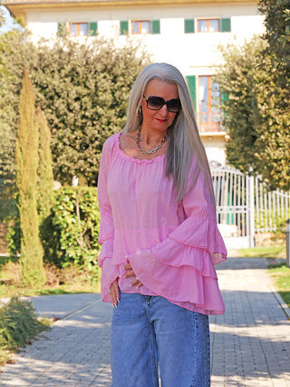 Person trägt das OFF#DLY Blusenshirt Nena pink mit voluminösen Volantärmeln. Es fällt locker und leicht, ideal für einen sommerlichen Look, kombiniert mit blauer Jeans und goldener Halskette.
