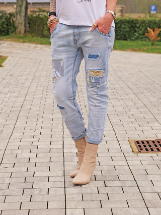 OFF#DLYDenim Destroy Camo lightblue Jeans mit zerrissenem Look und Camouflage-Patches, getragen mit nude-tan Boots. Sichtbare goldene Armbänder betonen den lässigen, trendigen Stil.