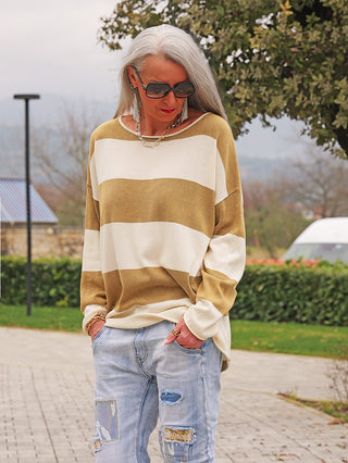 Person trägt OFF#DLY Pulli Ancona beige/khaki mit breiten Streifen und lockerem Schnitt, kombiniert mit hellen, zerrissenen Jeans und Accessoires, stehend auf einem gepflasterten Weg.