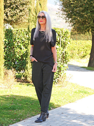 Frau trägt die OFF#DLY Denim black/black Lola, eine locker geschnittene, zeitlose schwarze Jeans mit hohem Tragekomfort, kombiniert mit schwarzen Stiefeln und silbernen Accessoires, in einem Garten.