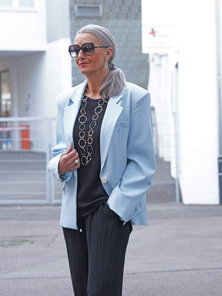 Die reife Frau trägt den OFF#DLY Blazer Venezia lightblue, ein stilvoller, leicht oversized Blazer mit klassischem Revers und lockerer Passform, kombiniert mit schwarzen, plissierten Hosen für einen minimalistischen Look.
