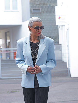 Mature Frau trägt den OFF#DLY Blazer Venezia lightblue, ein eleganter Oversize-Blazer mit Reverskragen, kombiniert mit schwarzem Top und strukturierten Hosen, gestylt mit auffälligen Accessoires.