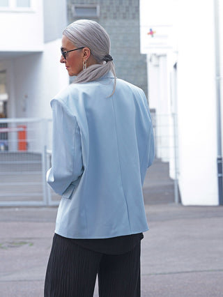 Person mit silbergrauem Haar im niedrigen Pferdeschwanz, trägt den OFF#DLY Blazer Venezia lightblue. Der Oversize-Blazer verleiht eine moderne Silhouette, kombiniert mit schwarzen Hosen und goldenen Ohrringen.