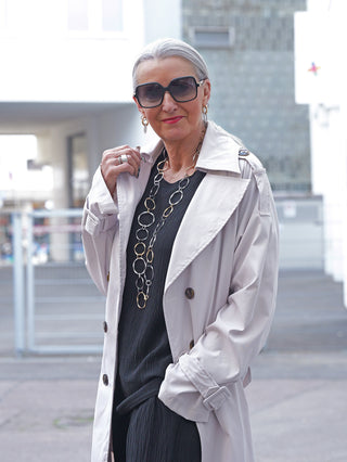 Ältere Frau trägt OFF#DLY Trenchcoat hellgrau über schwarzem Outfit, kombiniert mit großer Sonnenbrille und Kettenhalskette, verkörpert klassische Eleganz und modernen Stil in urbaner Umgebung.
