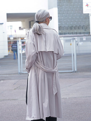 Person von hinten, trägt OFF#DLY Trenchcoat hellgrau mit locker gebundenem Gürtel. Der Mantel hat eine klassische Silhouette, doppelte Knopfreihe und Trench-Kragen, ideal für eleganten, modernen Stil.