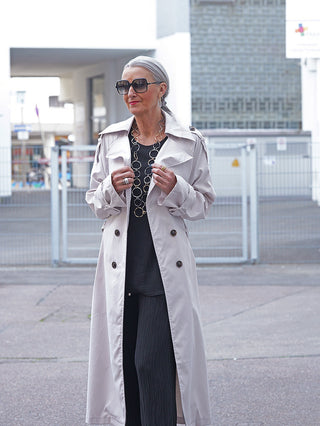 Ältere Frau in OFF#DLY Trenchcoat hellgrau, kombiniert mit schwarzem Outfit, Sonnenbrille und Schmuck, steht auf städtischem Gehweg. Elegante und moderne Silhouette wird durch den lockeren Fall des Mantels betont.