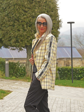 Ältere Frau trägt den OFF#DLY Blazer min yellow: Ein-Knopf-Design mit gelbem Karomuster und sportivem Logo-Streifen am Ärmel, kombiniert mit lockeren schwarzen Hosen.