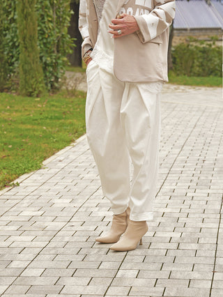 Person trägt OFF#DLY Hose Ida white, eine lockere, plissierte Bundfaltenhose aus weichem Material, kombiniert mit beigefarbenen Stiefeletten. Elegante Silhouette mit Blazer und Spitzenoberteil.
