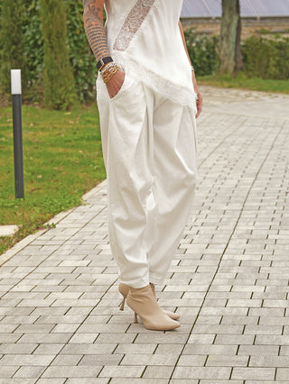 Person in weißem Outfit trägt die OFF#DLY Hose Ida, eine locker fallende Bundfaltenhose, kombiniert mit nudefarbenen Stiefeletten. Goldene Armbänder und ein tätowierter Unterarm sind sichtbar.