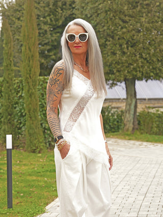 Frau mit silbergrauen Haaren trägt das OFF#DLY Top Hedy white, ein elegantes, weißes Spitzen-Top mit verstellbaren Trägern und fließendem Stoff, kombiniert mit weißen Hosen.
