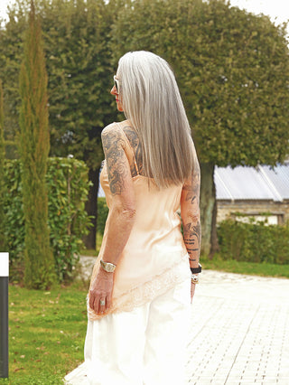 Person von hinten auf Gartenweg, trägt das OFF#DLY Top Hedy orange mit Spitzenrand und verstellbaren Trägern. Die Arme sind tätowiert.