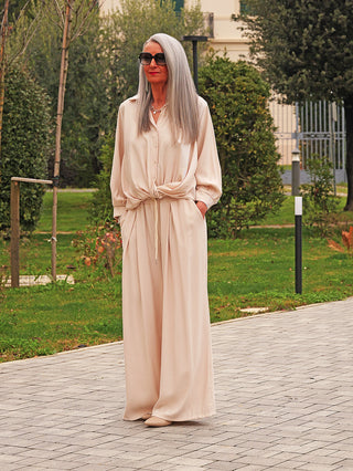 Frau trägt die OFF#DLY Hose Bari vanille mit weitem Schnitt und Bundfalten auf einem Gartenweg. Sie kombiniert sie mit einer beige Bluse und passenden Flats, was einen eleganten Look schafft.