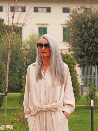 Frau im Park trägt die OFF#DLY Bluse Rom vanille, locker gebunden an der Taille, mit langen Ärmeln und dezenter Knopfleiste, kombiniert mit passender Hose für einen eleganten Look.