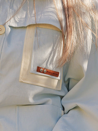 Nahaufnahme der OFF#DLY Jacke Mira lightblue mit zweifarbigem Design, sichtbarer Brusttaschenklappe mit Lederetikett, großem Knopf und Sonnenlicht, das weiche Schatten auf das strukturierte Baumwollmaterial wirft.