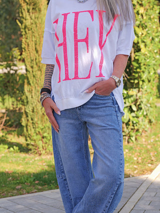 Person trägt das OFF#DLY Tee HEY white, ein lässiges, oversized T-Shirt mit pinkem HEY-Print, kombiniert mit hellblauen Jeans. Armbänder links, Uhr rechts, Tätowierung sichtbar.