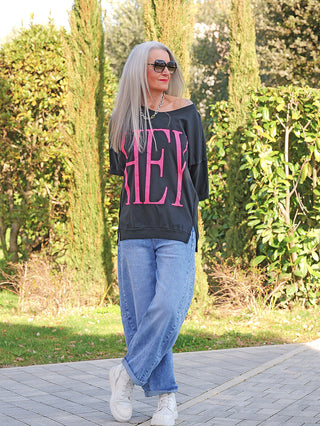 Person mit silbernem Haar trägt das OFF#DLY Tee HEY black, ein oversize schwarzes T-Shirt mit pinkem „HEY“-Aufdruck, kombiniert mit blauen Jeans und weißen Sneakers, in einem parkähnlichen Umfeld.
